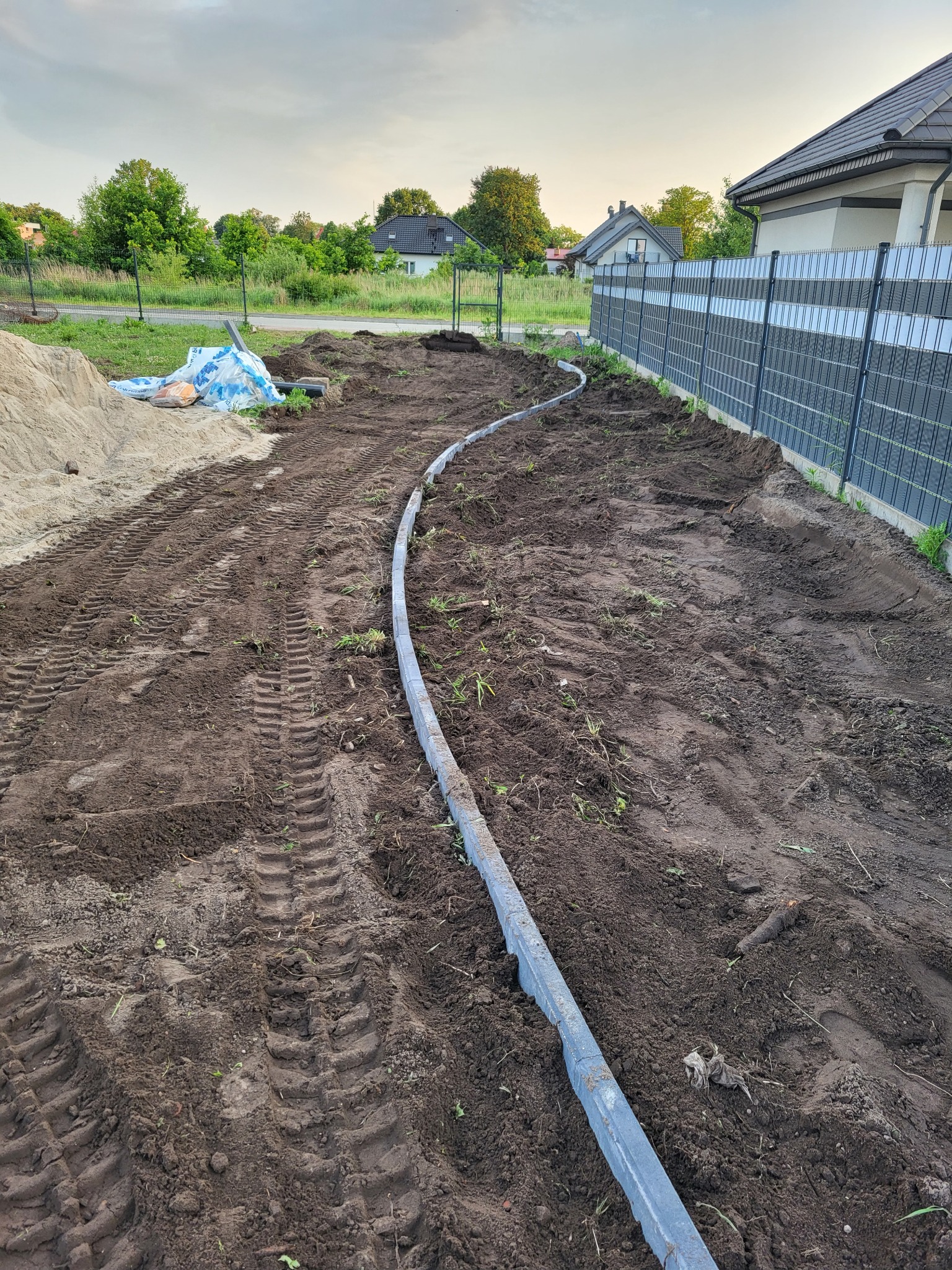 Przygotowanie terenu pod ścieżkę w ogrodzie z ułożonymi obrzeżami, widoczne ślady opon i pryzma piasku w tle.