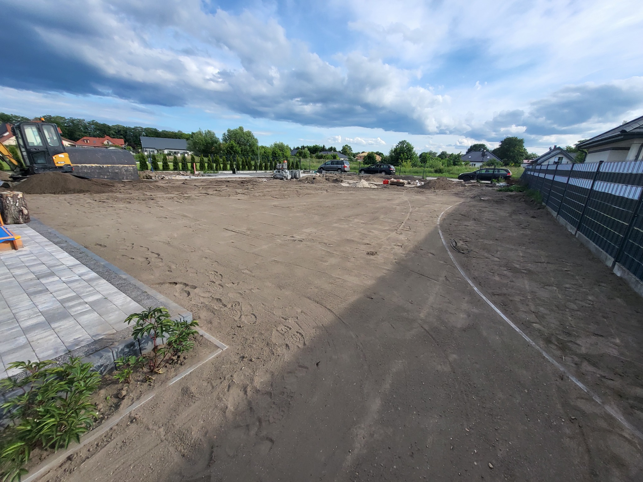 Przygotowany teren pod przyszły ogród z widoczną kostką brukową, świeżo posadzone rośliny, koparka w tle i nowo postawione ogrodzenie panelowe.