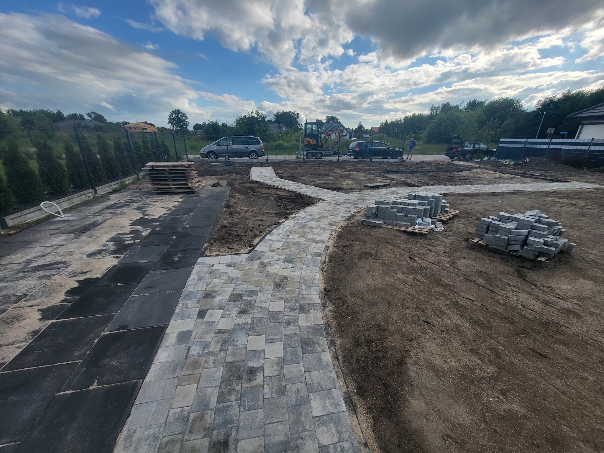 W trakcie budowy ścieżki z kostki brukowej w ogrodzie, widoczne ułożone fragmenty, palety z kostką, mini koparka i samochody w tle.