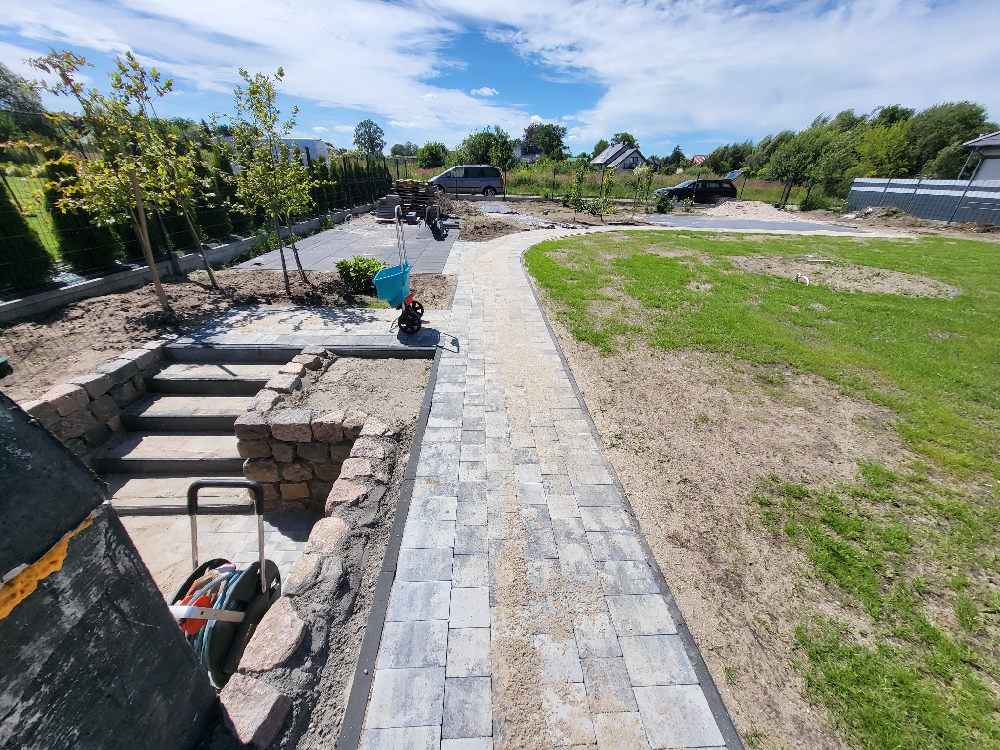Nowa ścieżka z kostki brukowej w ogrodzie, z widocznymi schodami z kamienia, trawnikiem i ogrodzeniem w tle, na etapie prac wykończeniowych.
