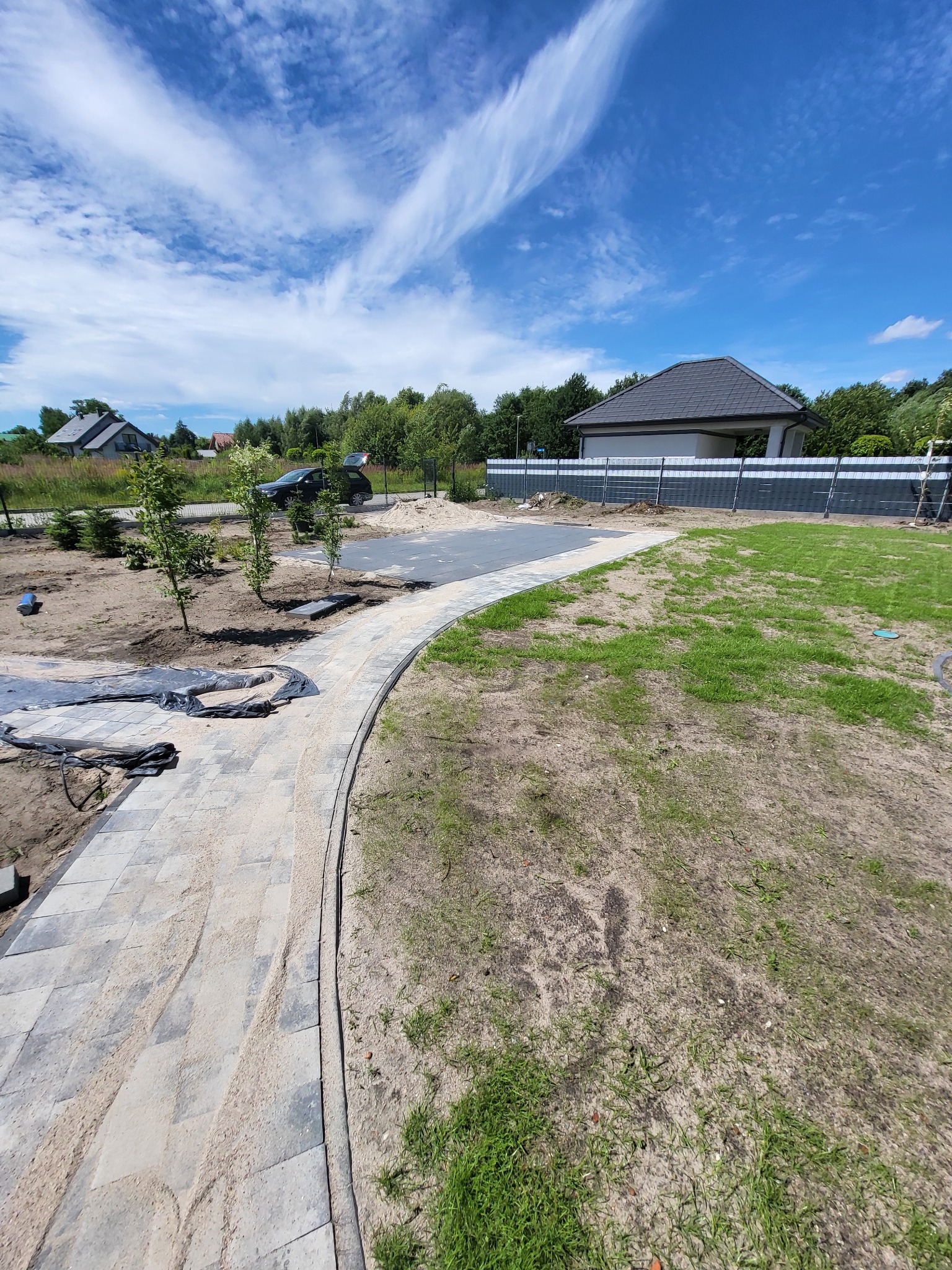 Krzywoliniowy chodnik z kostki brukowej w trakcie budowy, podsypany piaskiem, prowadzący do utwardzonego placu parkingowego, w tle dom jednorodzinny i błękitne niebo z chmurami.