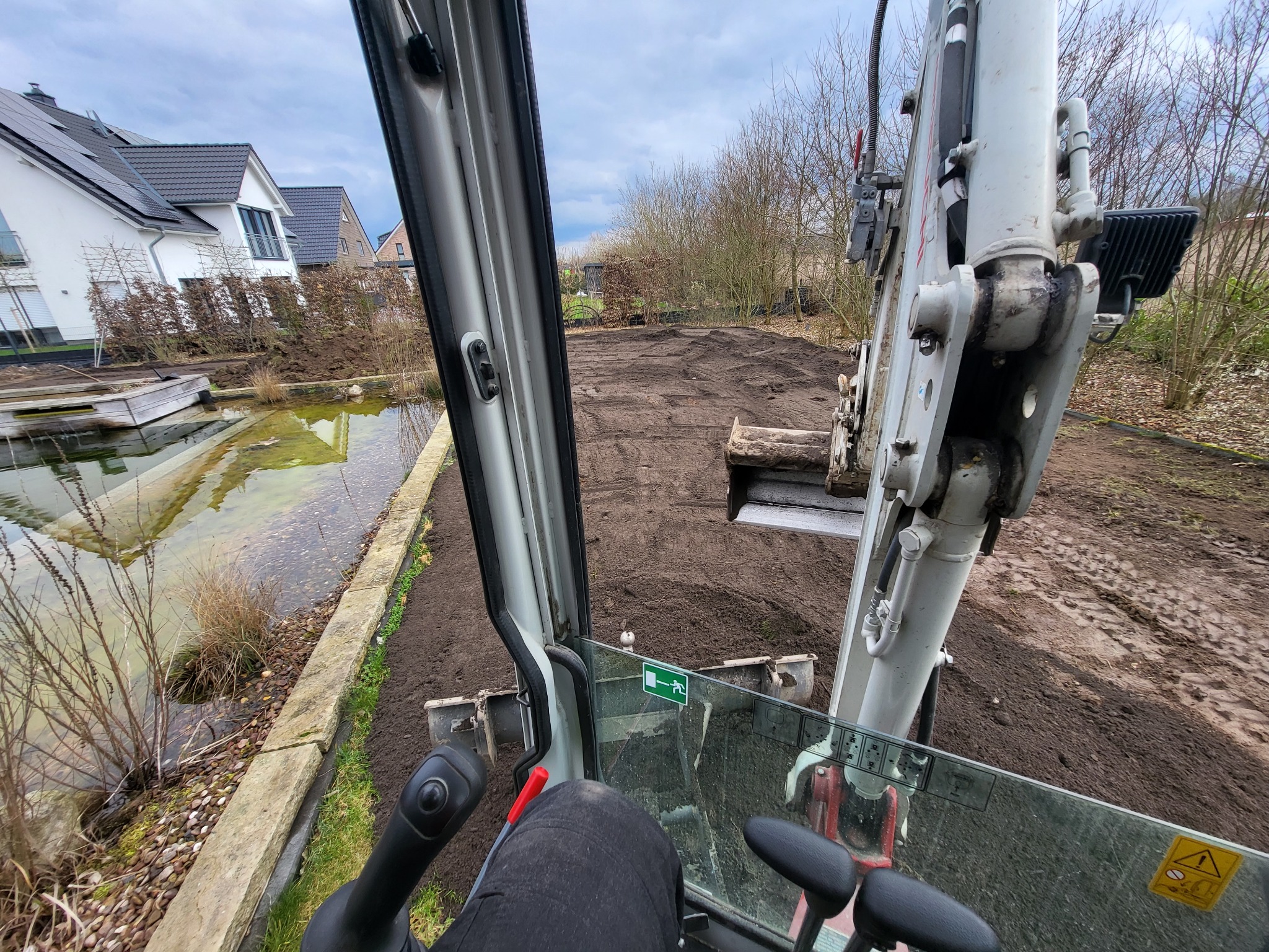 Widok z kabiny minikoparki na wyrównywany teren pod przyszły ogród, z widocznym stawem w tle i domem z panelami słonecznymi po lewej stronie.