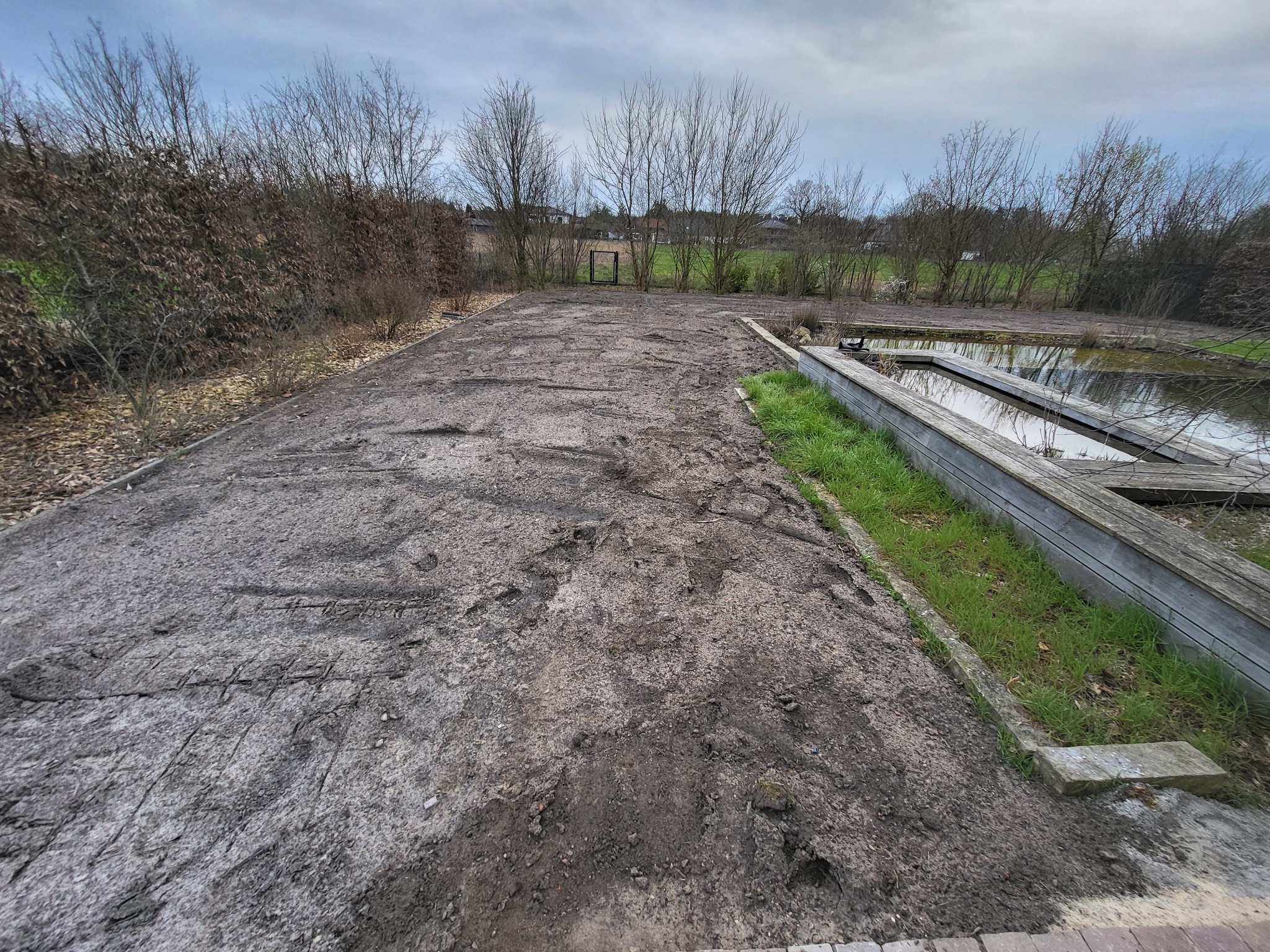 Przygotowany teren pod przyszły ogród z widocznymi śladami po pracach ziemnych, w tle drzewa bez liści i niewielki zbiornik wodny z drewnianą obudową.