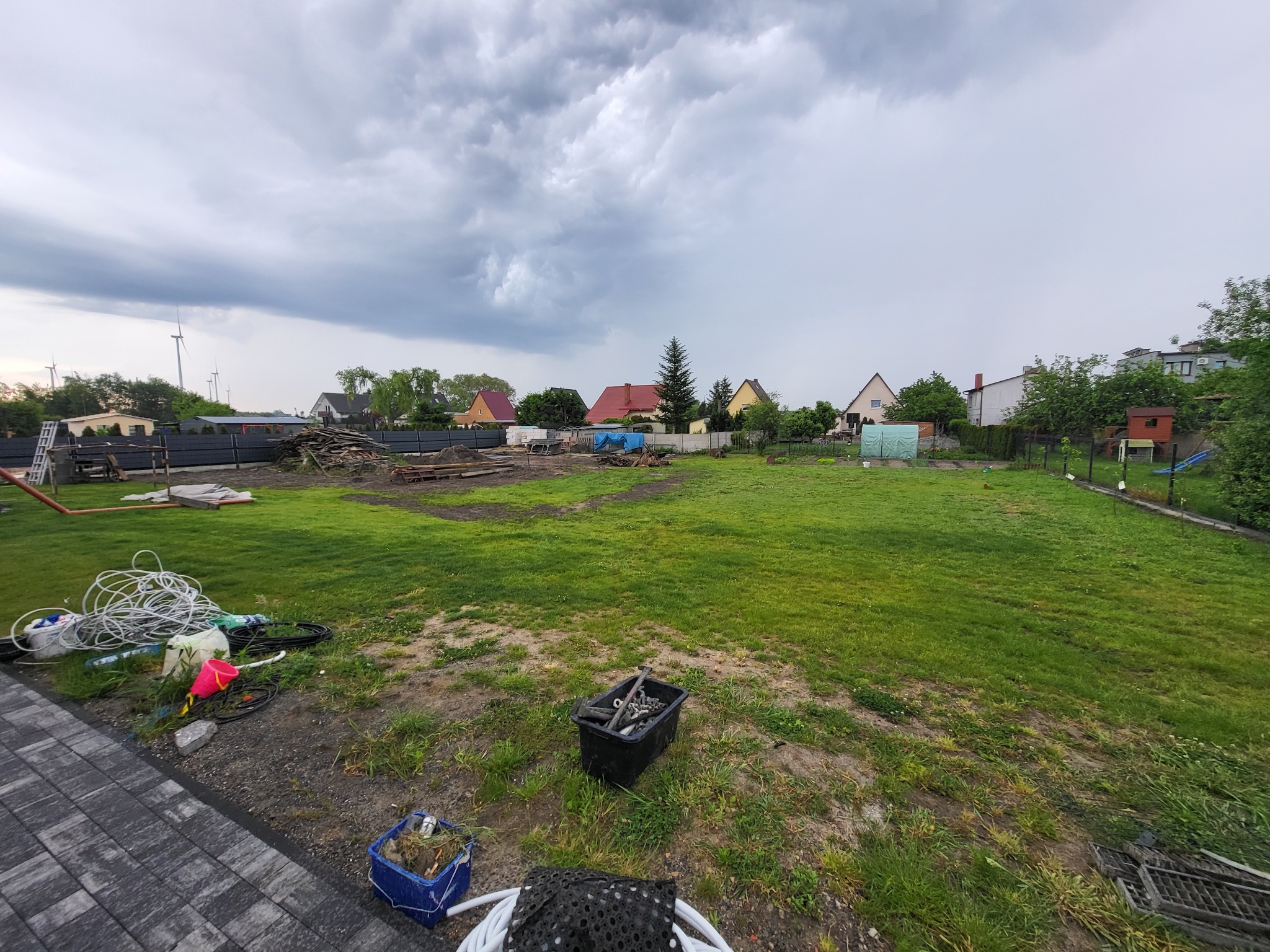 Przygotowanie terenu pod przyszły ogród: rozległy trawnik z widocznymi narzędziami i materiałami budowlanymi, w tle zabudowa jednorodzinna i pochmurne niebo.
