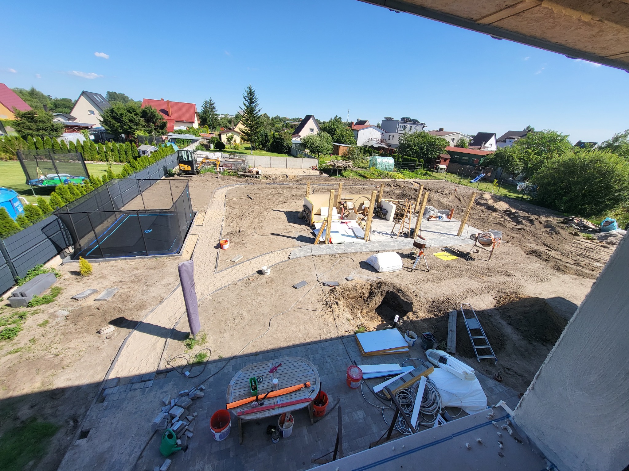 Widok z góry na plac budowy w ogrodzie: przygotowanie terenu, fundamenty pod konstrukcję drewnianą, wytyczone ścieżki, wykop, narzędzia, częściowo ułożona kostka brukowa, w tle domy i zieleń.