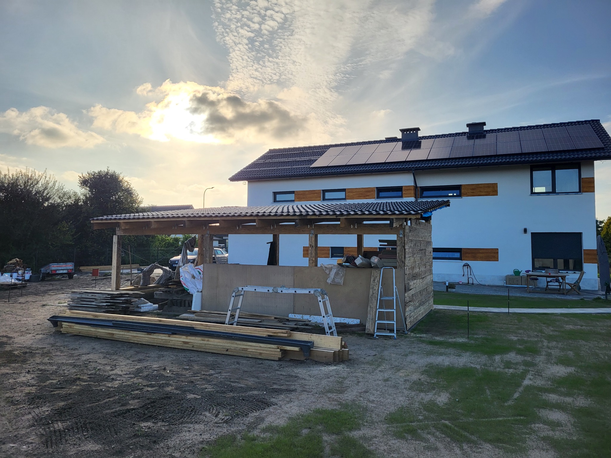 Budowa zadaszenia tarasu przy nowoczesnym domu z panelami słonecznymi, widoczne drewniane elementy konstrukcyjne, płyty i drabiny na placu budowy.