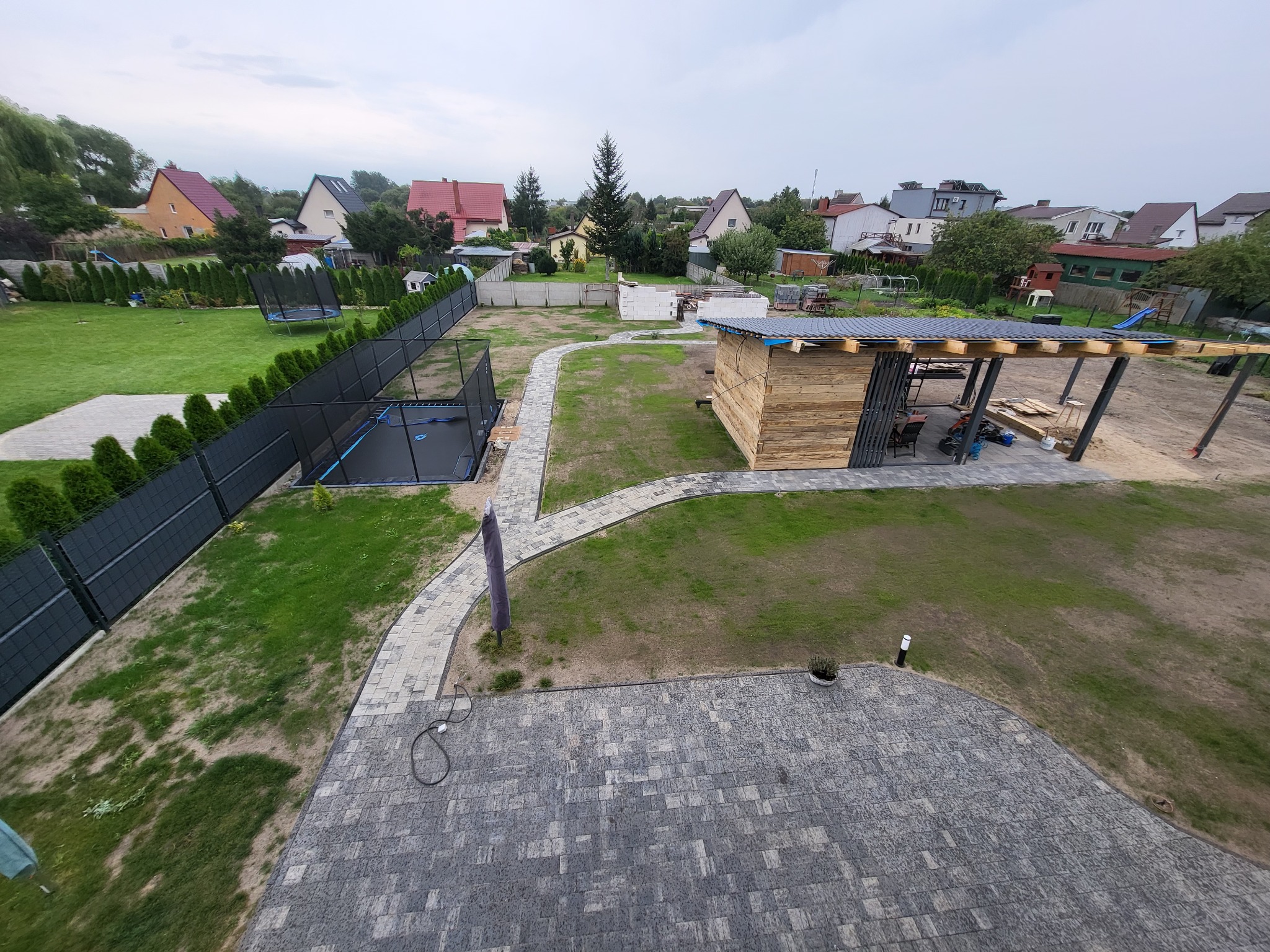 Widok z góry na ogród w trakcie realizacji: ułożona kostka brukowa, ścieżka z kostki, budowa drewnianej konstrukcji z zadaszeniem, czarne ogrodzenie panelowe, trampolina i domy w tle.