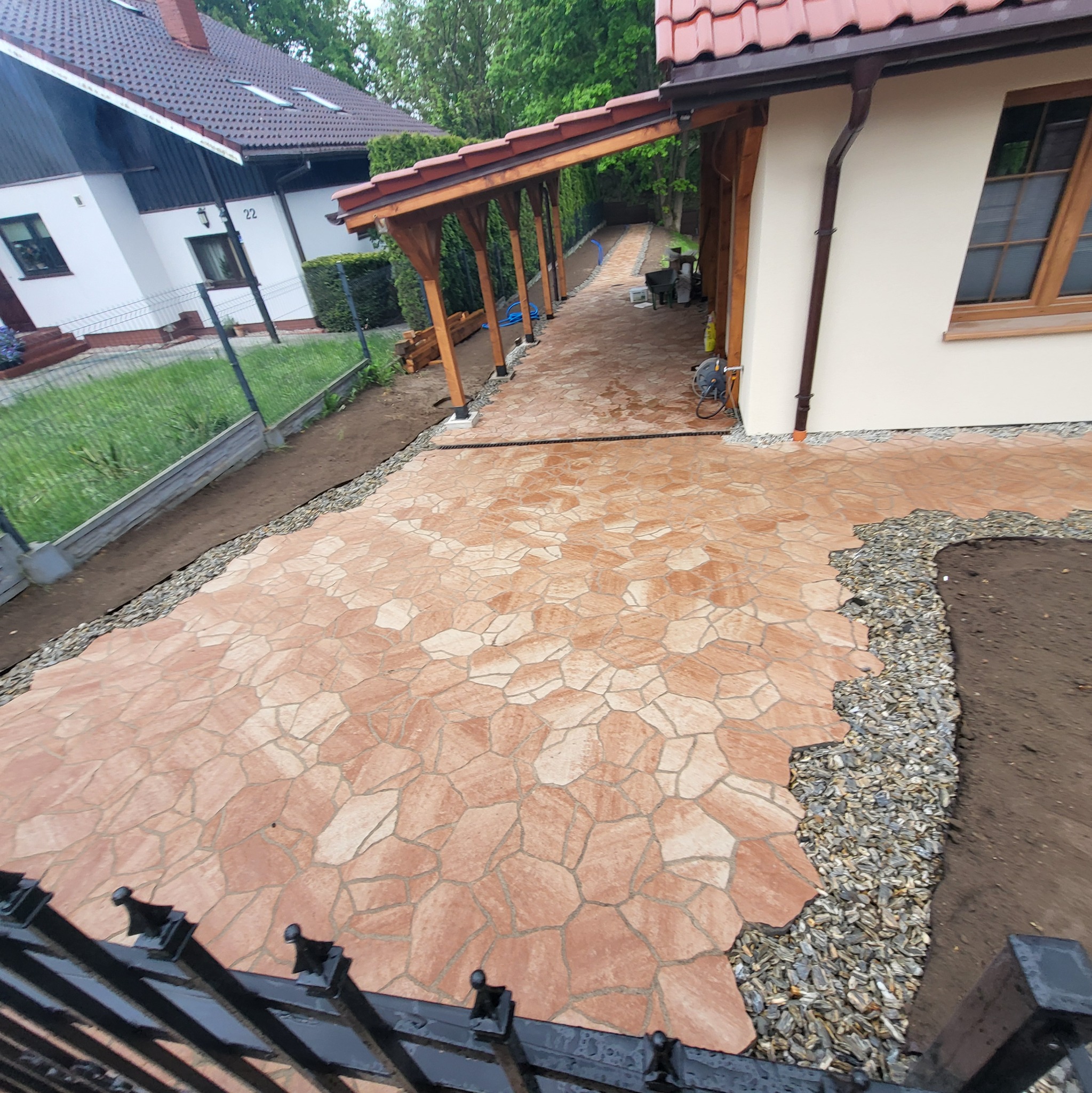 Wykończona ścieżka z nieregularnej kostki w odcieniach brązu prowadząca wzdłuż domu z drewnianą pergolą i rabatami z szarego żwiru. Fragment czarnego ogrodzenia.