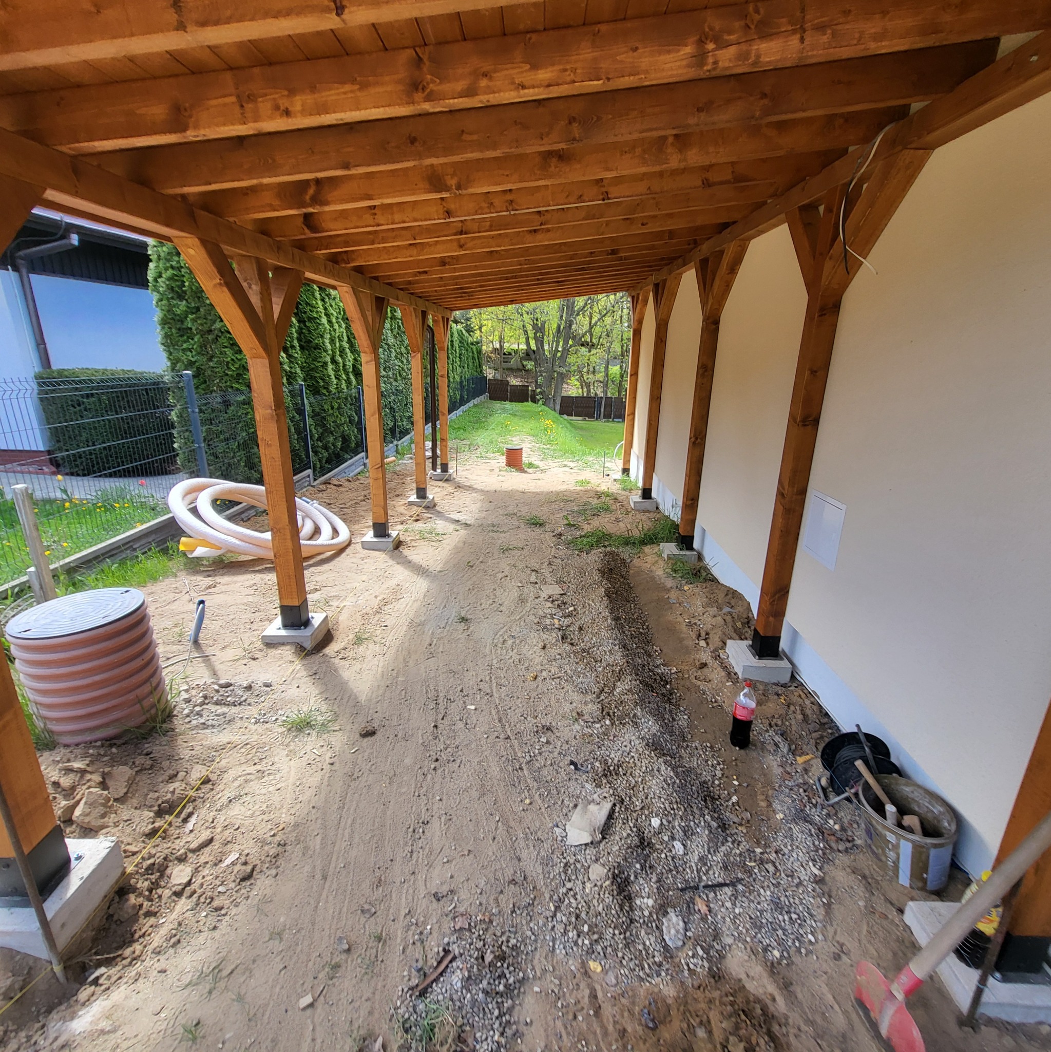 Drewniana konstrukcja zadaszenia w trakcie budowy, widoczne słupy na betonowych fundamentach, teren przygotowany pod dalsze prace, narzędzia budowlane.