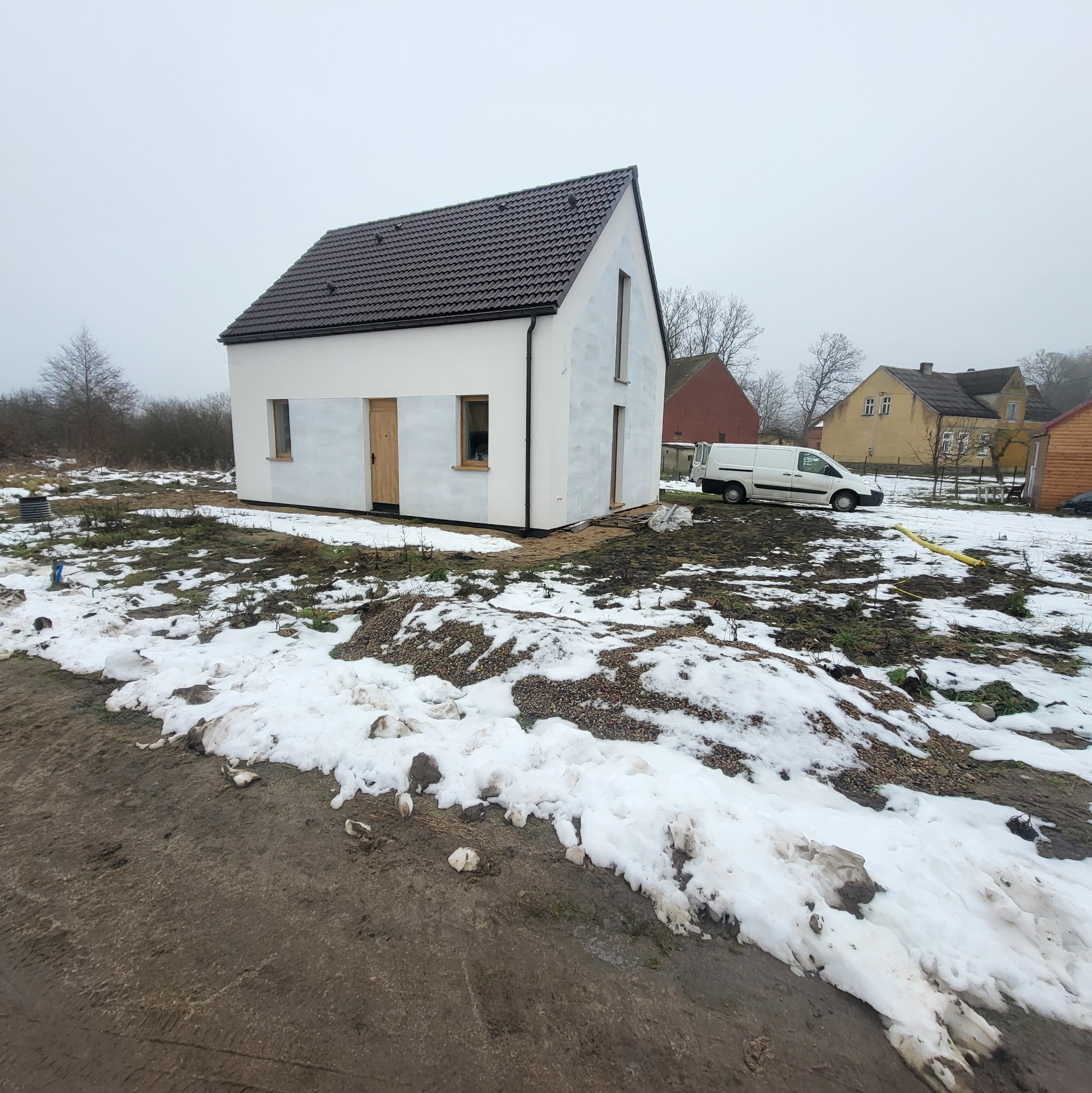 Surowy, biały dom z brązowym dachem w zimowym krajobrazie. Widoczny samochód dostawczy i ślady prac budowlanych. Minimalistyczna architektura w trakcie wykończenia.