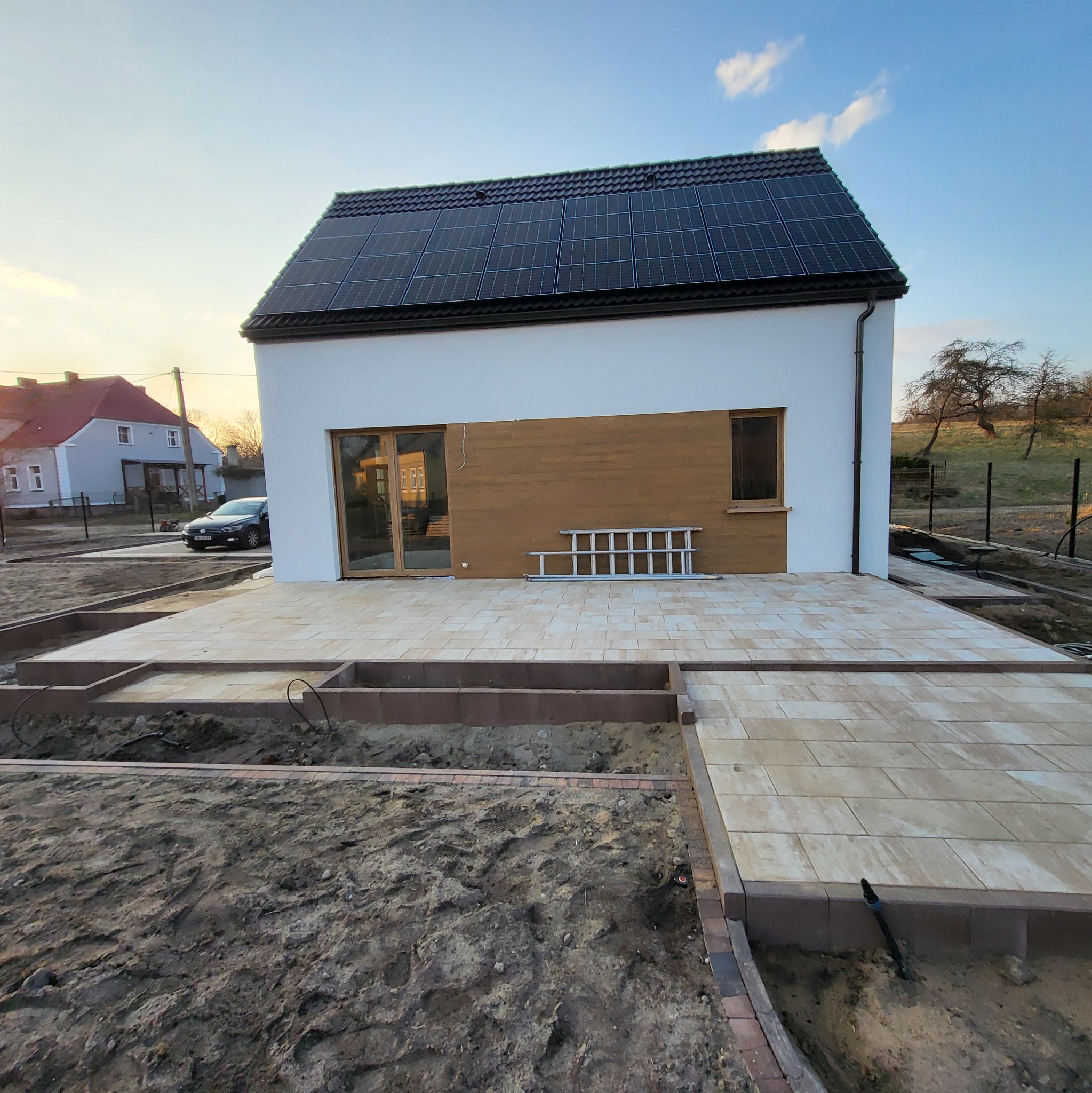 Nowoczesny dom z panelami słonecznymi i częściowo ułożonym tarasem z kostki. Widoczne przygotowane podłoże pod dalszą część tarasu. Drabina oparta o ścianę.