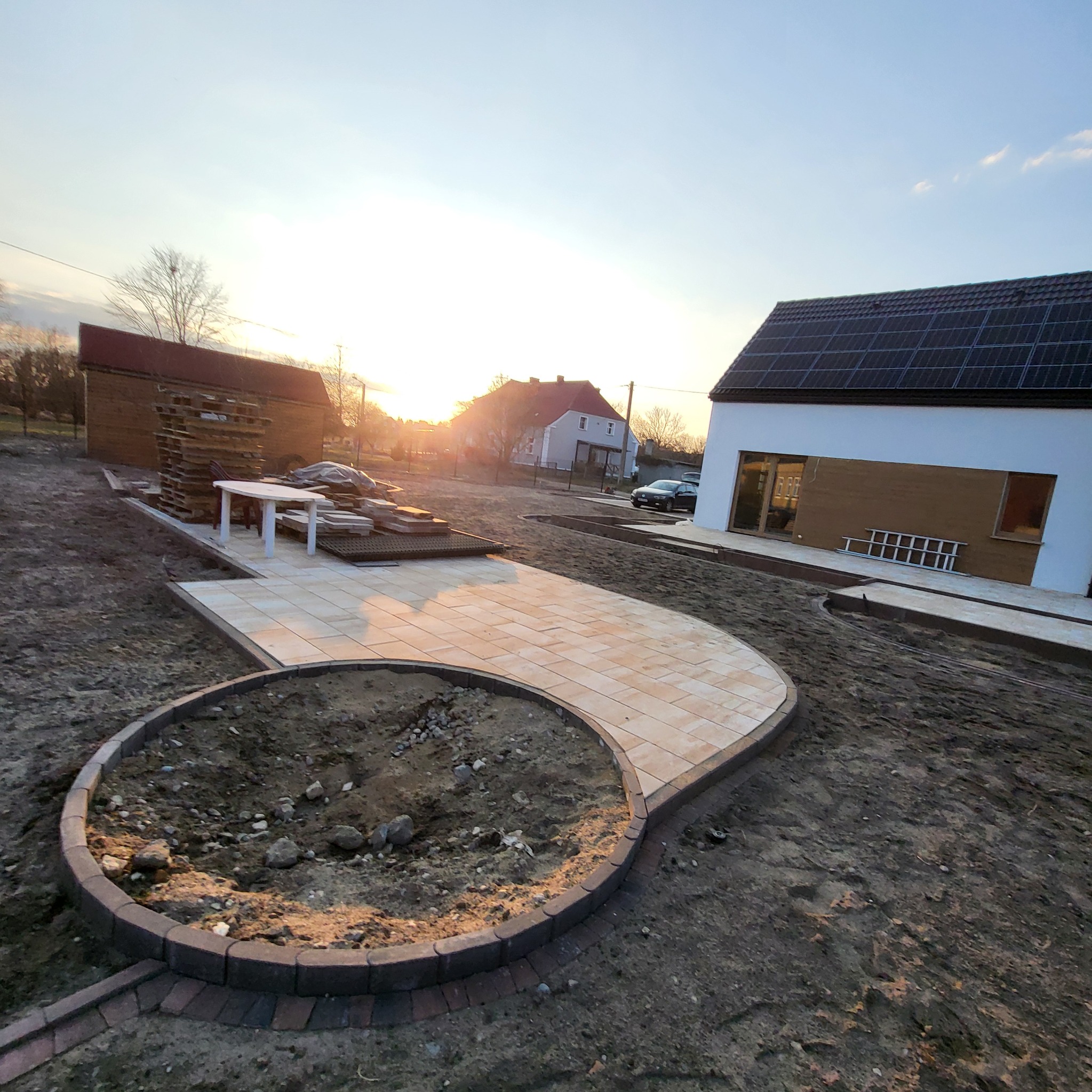 Układanie kostki brukowej w ogrodzie, widok z góry na ścieżkę i rabatę, w tle dom z panelami słonecznymi, zachód słońca.