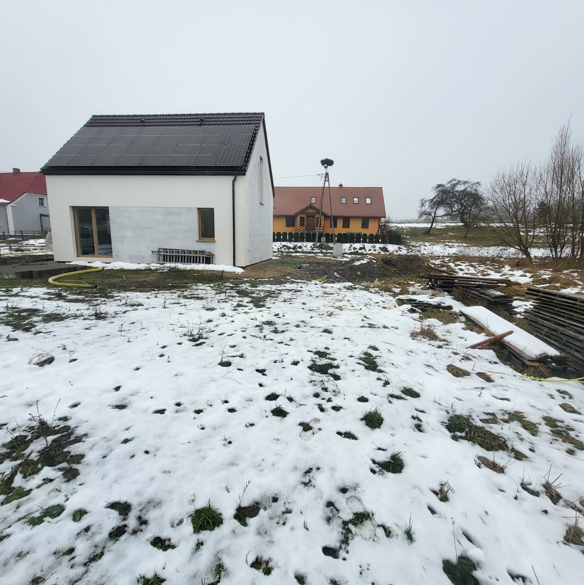 Nowoczesny dom z panelami słonecznymi na dachu, w zimowym krajobrazie. Widoczne elementy ogrodu w trakcie realizacji i sąsiedni budynek z gniazdem bocianim.