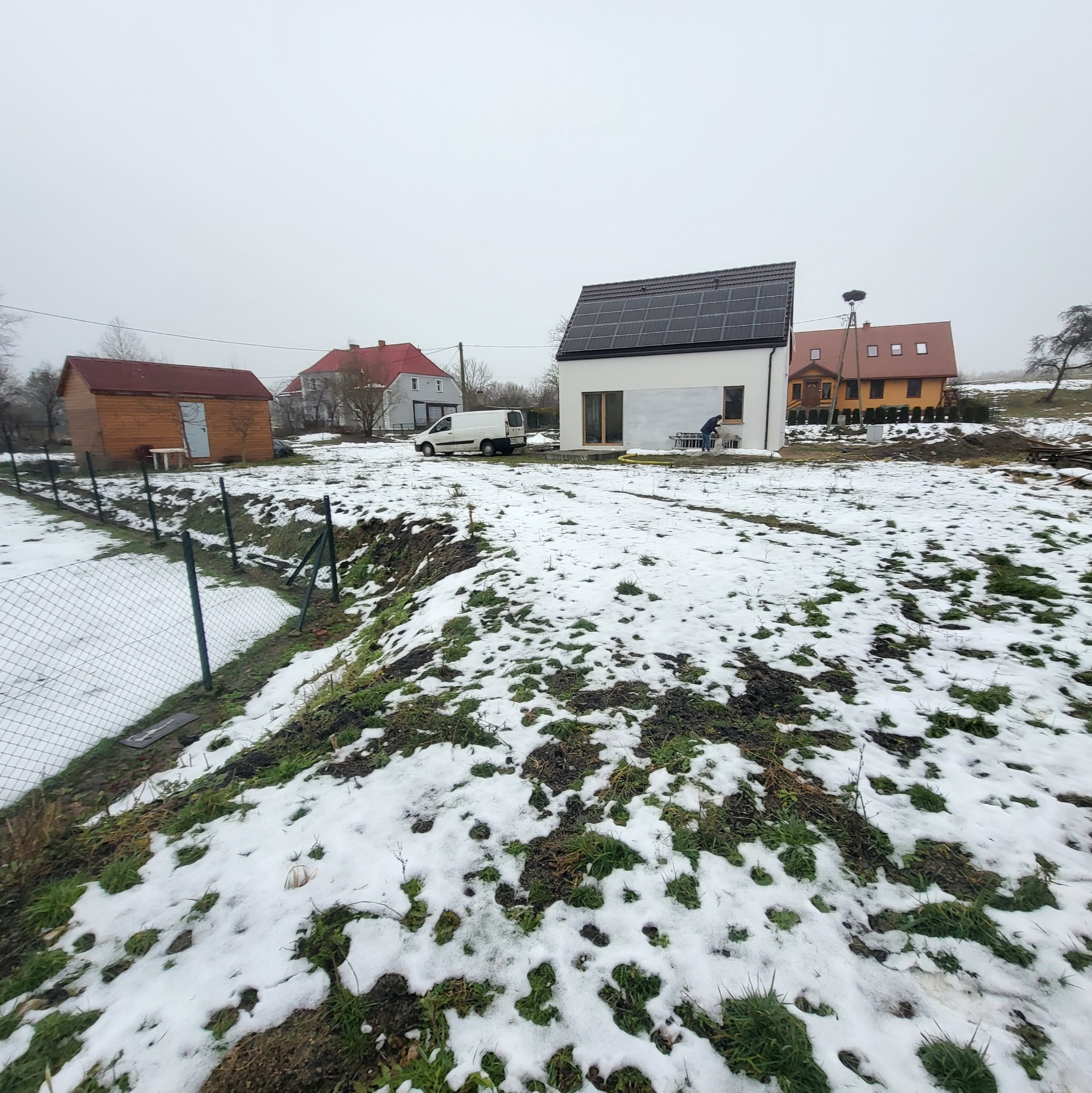 Nowoczesny dom z panelami słonecznymi na dachu, widok z zasypanego śniegiem ogrodu. W tle inne zabudowania i częściowo widoczny samochód dostawczy.