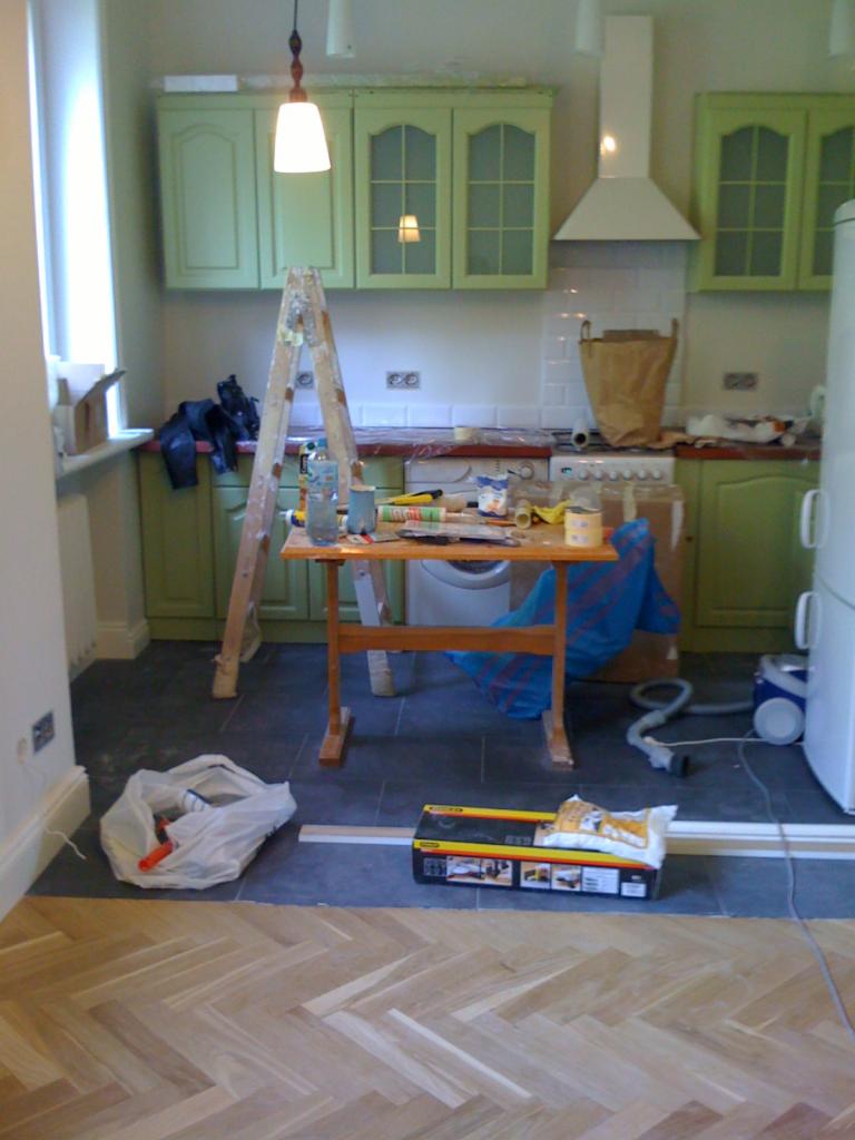 Kuchnia w trakcie remontu: parkiet ułożony do połowy, drabina oparta o szafki, narzędzia na stole. Widoczne materiały budowlane i niedokończona podłoga.