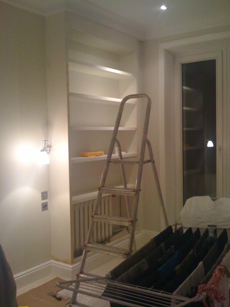 Biała zabudowa z półkami wnękowymi, drabina aluminiowa, suszarka z praniem w pokoju podczas wykańczania. Widoczne listwy przypodłogowe i gniazdka elektryczne.