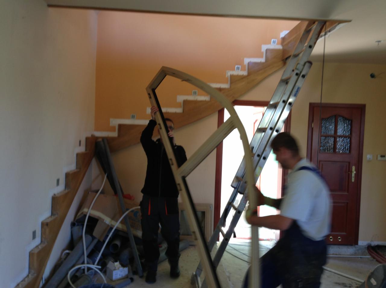 Montaż ramy okiennej w dwupoziomowym wnętrzu z drewnianymi schodami i drabiną. Widoczni dwaj pracownicy w trakcie pracy. W tle drzwi i pomarańczowa ściana.