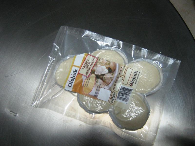 Pakowane krążki ciasta na pierogi marki Olejnik, widoczne przez przezroczyste opakowanie, leżące na metalowej powierzchni.