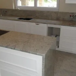 Wykończona kuchnia z białymi szafkami i blatem z jasnego granitu. Widoczna płyta indukcyjna i zlew. Okno z ceglanym widokiem na zewnątrz. Neutralne kolory ścian i podłogi.