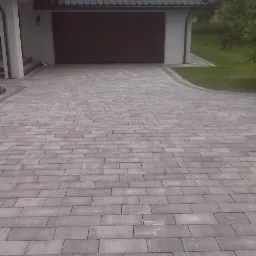 Szara kostka brukowa na podjeździe domu z garażem, widok z perspektywy. Estetyczne wykończenie, równa powierzchnia, obrzeża z kostki. Zieleń trawnika w tle.