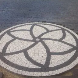 Okrągły wzór kwiatowy z jasnoszarej i ciemnoszarej kostki granitowej, wkomponowany w nawierzchnię z ciemnej kostki brukowej; widoczne niedokończone krawędzie i podsypka piaskowa.