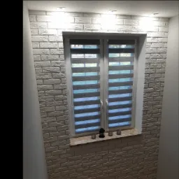 Ściana wewnętrzna wykończona białą cegłą dekoracyjną, okno z żaluzjami dzień-noc, oświetlenie punktowe sufitowe, dekoracje na parapecie.