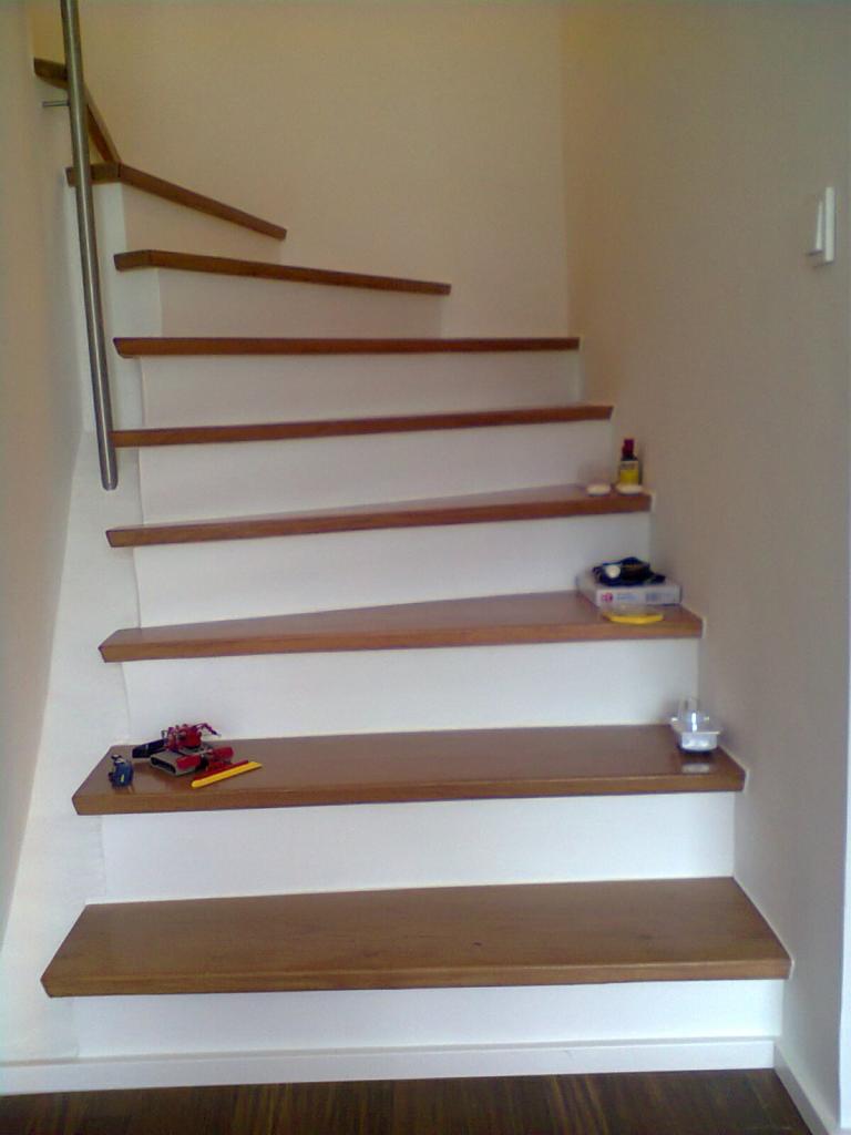 Schody drewniane z białymi podstopnicami, z metalową barierką po lewej stronie. Na stopniach leżą zabawki i inne przedmioty. Widok z dołu.