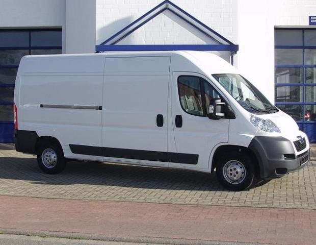 Biały, dostawczy bus na kostce brukowej, zaparkowany przed budynkiem z niebieskimi elementami architektonicznymi. Widok z boku, światło dzienne.