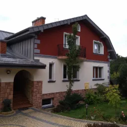 Dwukondygnacyjny dom jednorodzinny z czerwono-białą elewacją, szarą blachą na dachu, kamiennym podjazdem i bujną zielenią w ogrodzie.