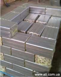 Ułożone na palecie brykiety drzewne Pini&Kay o prostokątnym przekroju, widoczne ślady owijki, słabe oświetlenie.