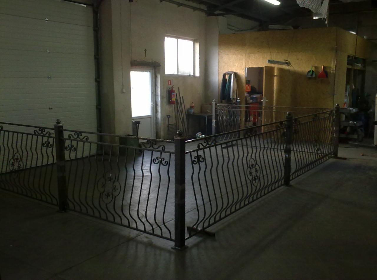 Metalowa balustrada z dekoracyjnymi elementami w kształcie kwiatów, zainstalowana w hali produkcyjnej, widoczna brama garażowa i okno.