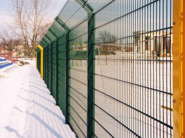 Ochronne ogrodzenie panelowe z siatki z elementami w kolorze zielonym i żółtym, otoczone śniegiem, z budynkiem w tle.