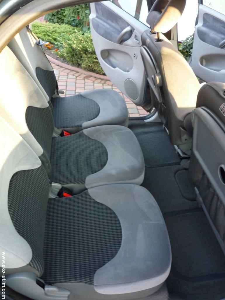 Wnętrze samochodu z widocznymi tylnymi siedzeniami z szarą tapicerką i elementami z tworzywa. Otwarta tylna klapa i widoczny fragment kostki brukowej na zewnątrz.