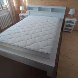 Białe łóżko z zagłówkiem i materacem w pokoju z drewnianą boazerią, obok szafki nocne z lampkami i krzesło przy grzejniku.