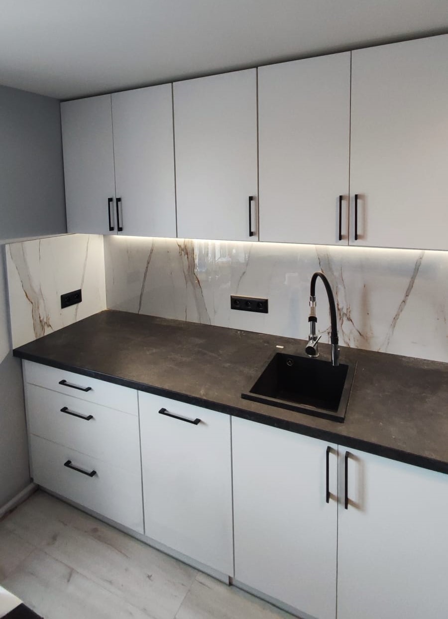 Nowoczesna kuchnia z białymi, lakierowanymi szafkami, czarnym blatem i marmurowym backsplashiem z podświetleniem LED. Minimalistyczne, czarne uchwyty i bateria.