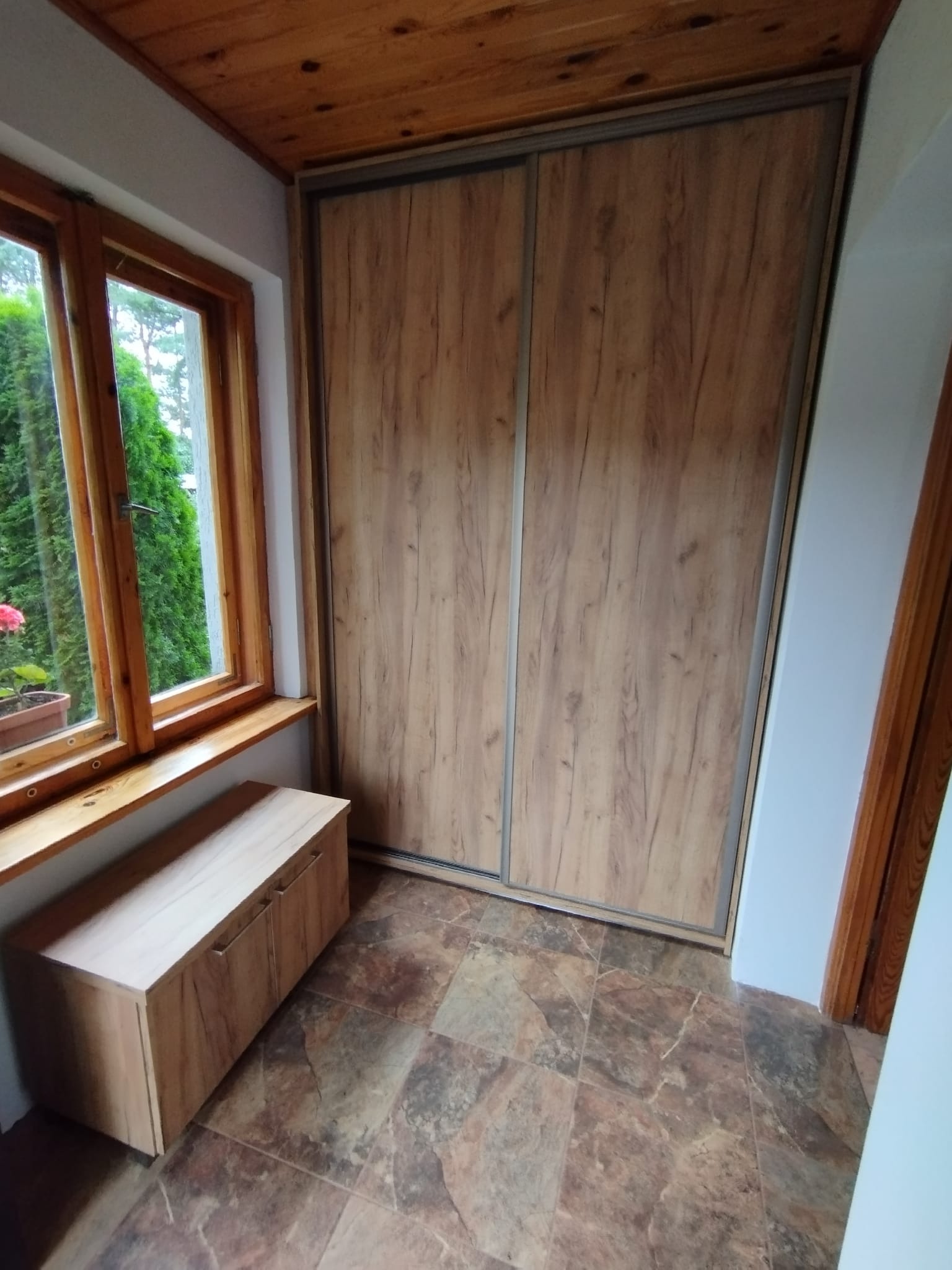 Wnęka z szafą przesuwną o drewnopodobnym wykończeniu, dopasowaną do drewnianej boazerii sufitowej i ramy okiennej, z dodatkową niską szafką pod oknem.