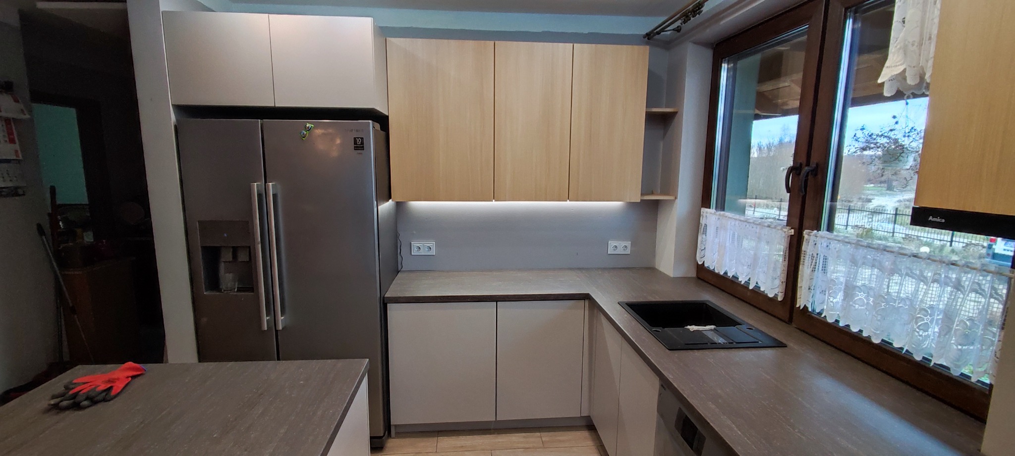 Nowoczesna kuchnia z szarymi i drewnianymi szafkami, oświetleniem LED pod szafkami wiszącymi i dużym oknem z koronkową firanką. Na blacie leżą czerwone rękawice robocze.