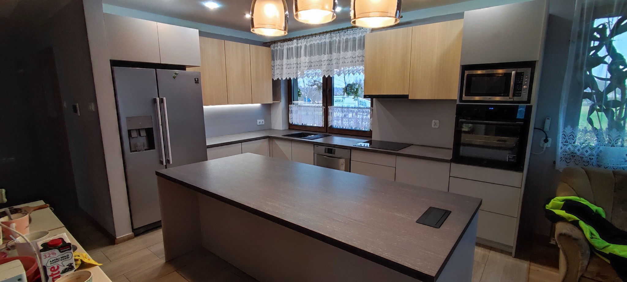 Nowoczesna kuchnia z wyspą, jasne fronty szafek górnych i szare dolne, stalowa lodówka z dystrybutorem wody, okno z koronkową firanką.