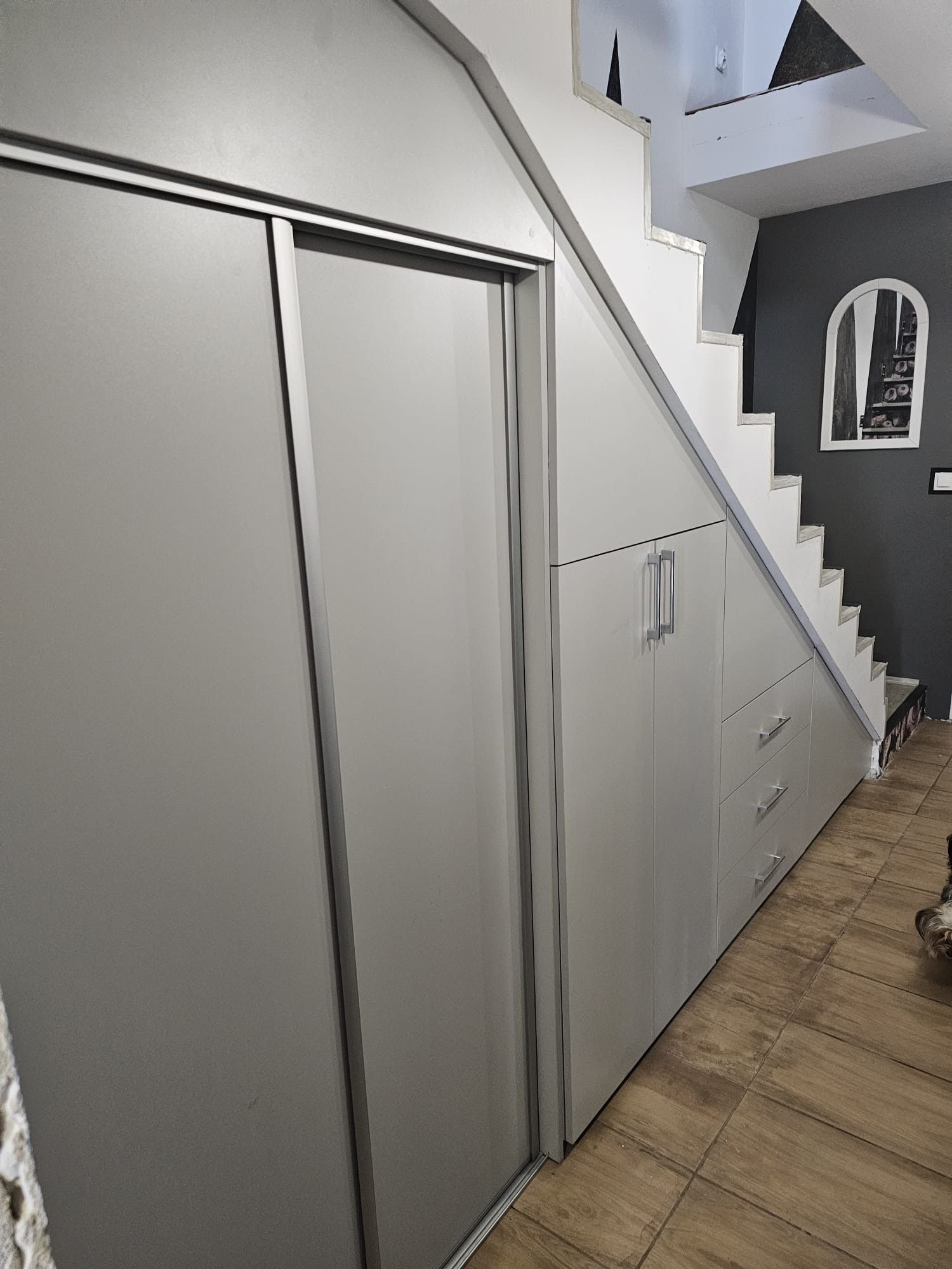 Szara szafa wnękowa ze srebrnymi uchwytami i przesuwnymi drzwiami, zabudowana pod schodami w mieszkaniu z drewnopodobną podłogą i szarą ścianą z dekoracyjnym lustrem.