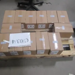 Paleta z kartonami, oznaczone białą taśmą i numerem C/03/4. Widoczny wózek widłowy i fragment krzesła. Transport i logistyka.