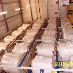 Magazyn z paletami, na których znajdują się duże worki typu Big Bag, prawdopodobnie z materiałami budowlanymi. Widoczne osoby w fartuchach i kaskach ochronnych na górze regałów.