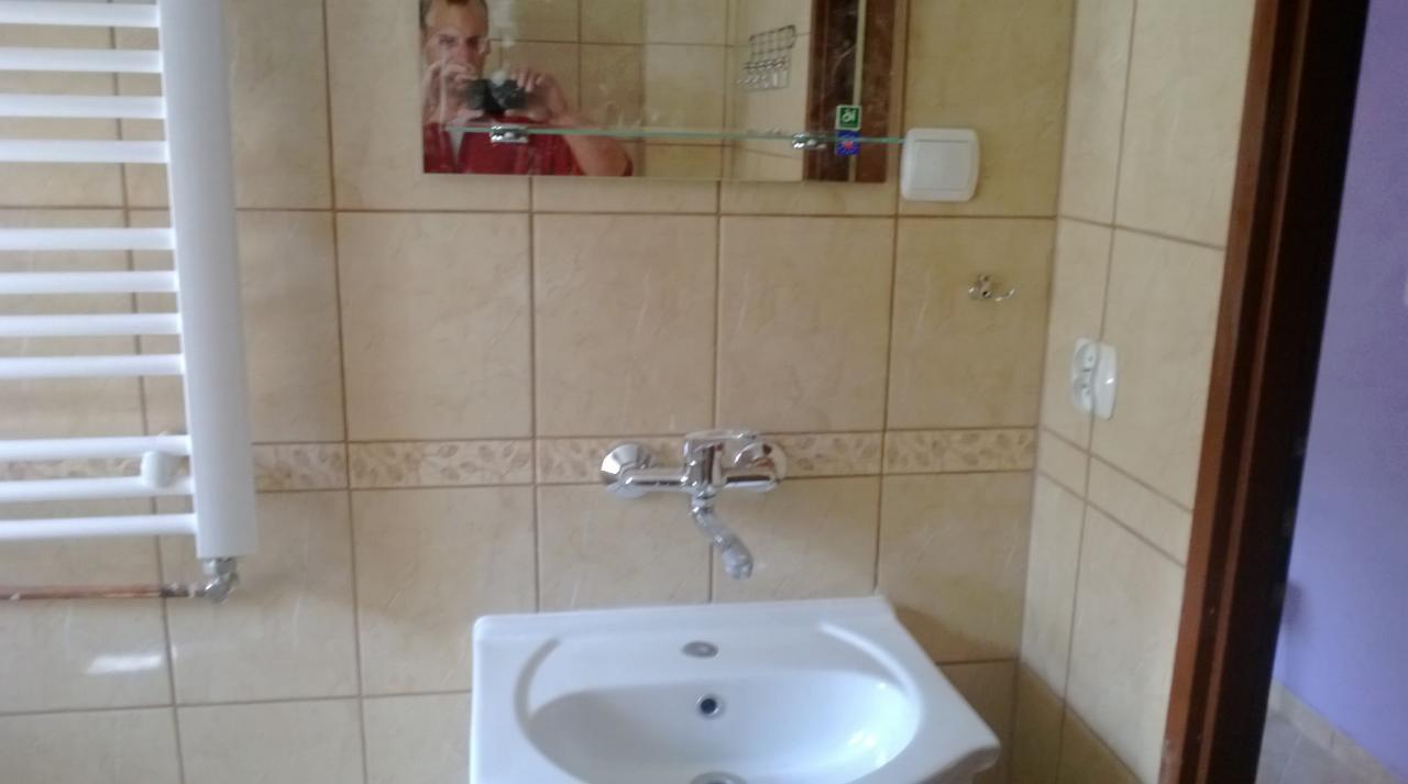 Beżowe płytki w łazience z lustrem odbijającym osobę, biała umywalka i grzejnik drabinkowy. Ujęcie z bliska na wykończoną łazienkę z elementami wyposażenia.