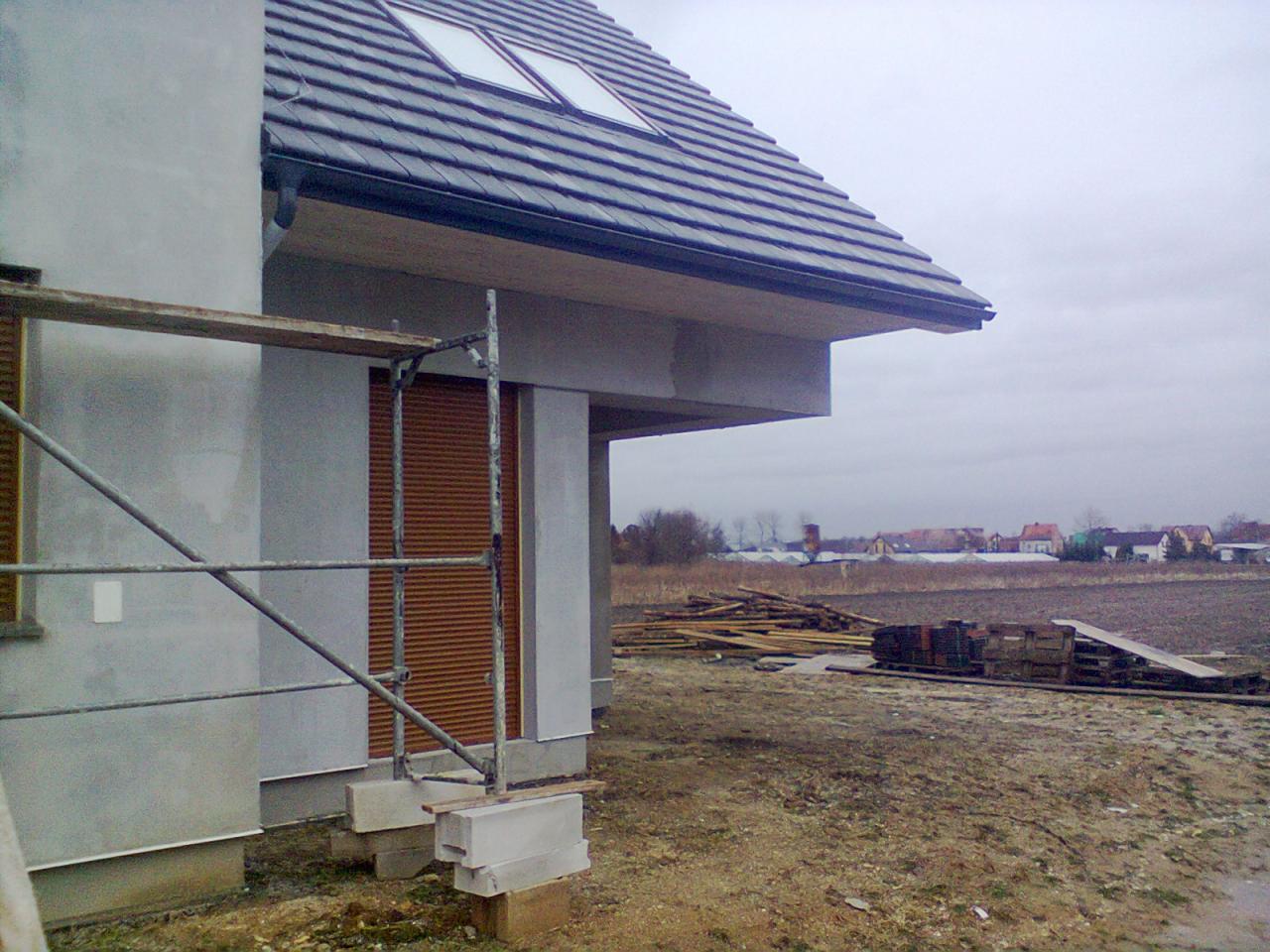 Dom w trakcie prac wykończeniowych: szare ściany, okna dachowe, rusztowanie, teren budowy. W tle zabudowa wiejska pod pochmurnym niebem.