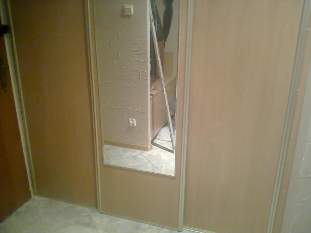 Beżowa szafa przesuwna z lustrem, jasne płytki podłogowe. Widoczny fragment ściany z teksturą imitującą cegłę i gniazdko elektryczne. Ujęcie z niskiego kąta.