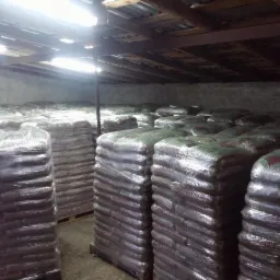 Magazyn z paletami worków, zabezpieczonych folią. Widoczne drewniane elementy konstrukcji dachu i oświetlenie magazynowe.