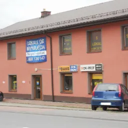 Budynek z oknami PCV, bramami roletowymi i drzwiami, z reklamami lokali do wynajęcia. Widoczne okna wystawowe z logo producentów.