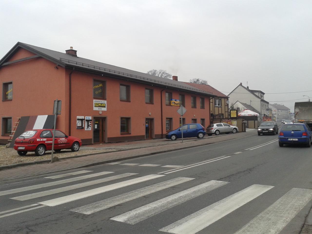 Budynek firmy Uni Blach z ekspozycją blach dachowych i zaparkowanym czerwonym autem reklamowym. Widok na ulicę z przejściem dla pieszych.