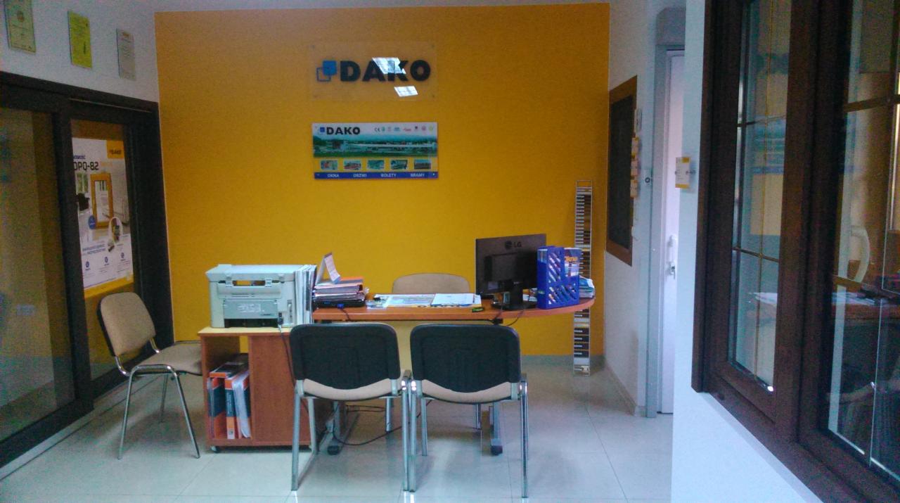Biuro firmy DAKO z ekspozycją okien i drzwi. Żółta ściana, biurko z komputerem i drukarką. W tle widoczny asortyment firmy.
