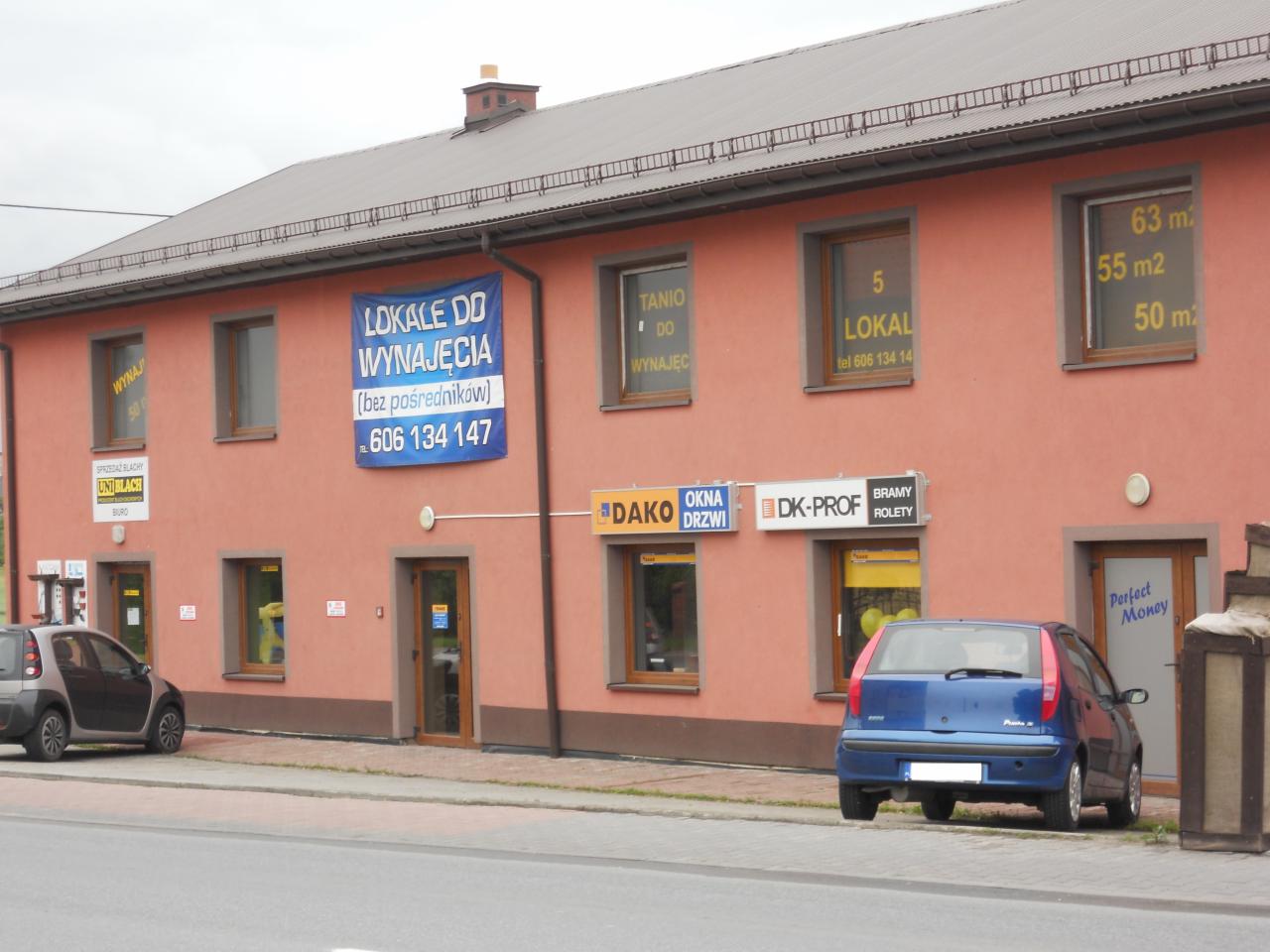 Budynek z oknami PCV, bramami roletowymi i drzwiami, z reklamami lokali do wynajęcia. Widoczne okna wystawowe z logo producentów.