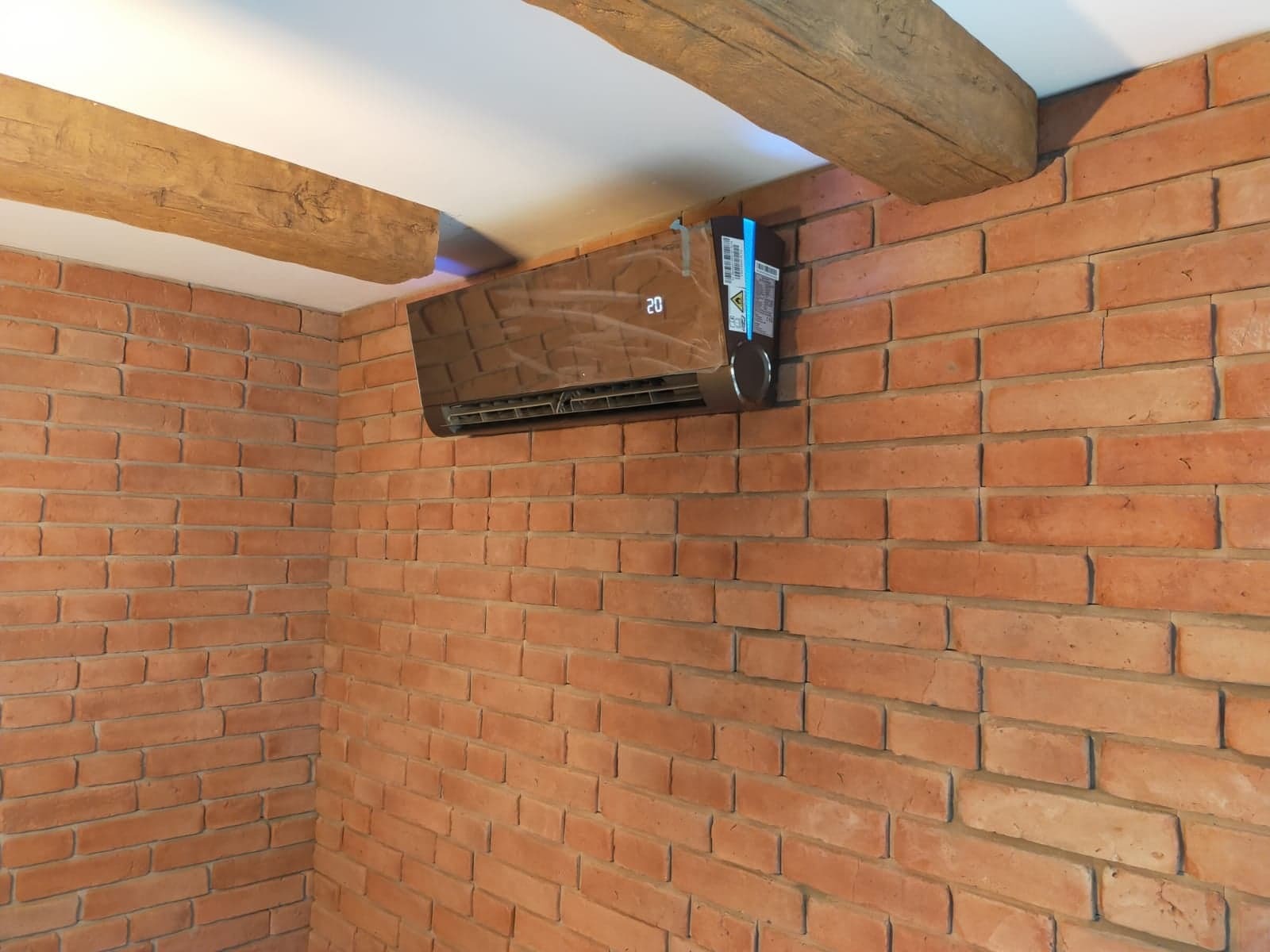 Brązowy klimatyzator ścienny z wyświetlaczem temperatury zamontowany na ścianie z cegły w rustykalnym wnętrzu z drewnianymi belkami stropowymi.